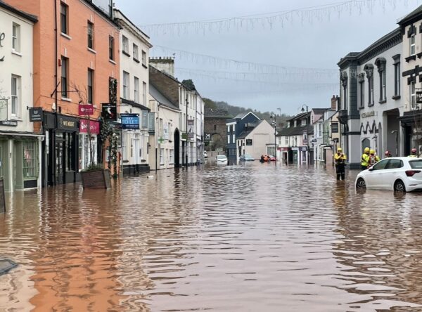 A town high street flooded with muddy water, partly submerging parked cars and shopfronts, as a few emergency workers stand in the water under a grey sky.